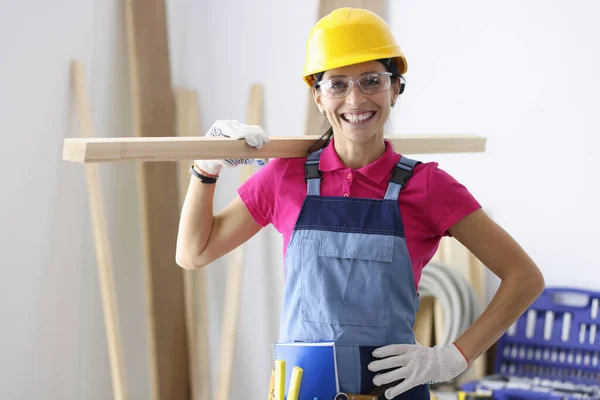 Žena ve stavební helmě a montérkách drží dřevěné prkno na rameni v dílně — Stock fotografie