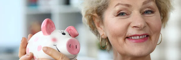 Happy elderly woman in apartment shows piggy bank