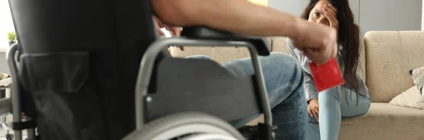Сумна молода жінка сидить на дивані навпроти чоловіка в інвалідному кріслі з презервативом в руці — стокове фото
