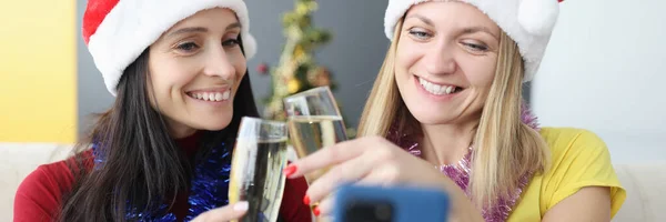 Duas mulheres rindo em chapéus de Papai Noel estão olhando para a tela do smartphone e segurando copos de champanhe — Fotografia de Stock