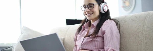 Mulher em fones de ouvido com laptop em seu colo está sentado no sofá e sorrindo. — Fotografia de Stock