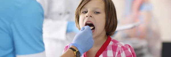 Médico en traje azul mira la garganta del niño con palo de madera. —  Fotos de Stock