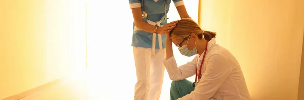 女性医師で保護医療マスクで座っています廊下で彼女の頭を下げ、彼女の同僚は彼女をサポート. — ストック写真