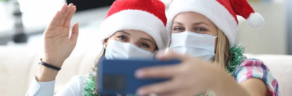 Duas mulheres amigas em máscaras protetoras e chapéus de Papai Noel vermelhos são fotografados no retrato do telefone — Fotografia de Stock