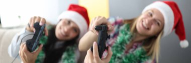 Kırmızı Noel Baba şapkalı iki kadın ellerinde joysticklerle kanepede oturmuş bilgisayar konsolu oynuyorlar.