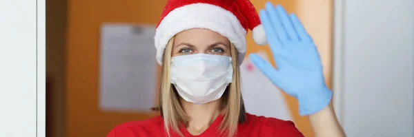 Vrouw postbode in kerstman hoed in medisch masker en rubber handschoenen houdt geschenk in haar hand en zwaait hallo — Stockfoto