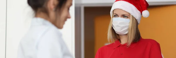 Koerier vrouw in rood kerstman hoed en beschermende medische masker op gezicht geeft klant om document en geschenk te ondertekenen — Stockfoto