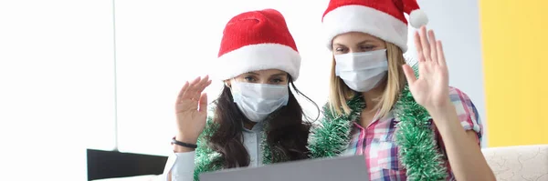 Dos mujeres con máscara protectora y sombrero de Santa Claus se sientan lado a lado y saludan. — Foto de Stock