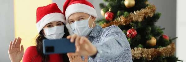 Hombre y mujer en sombreros de santa y máscaras protectoras en las caras están sosteniendo un teléfono móvil y saludando cerca del árbol de Navidad en casa — Foto de Stock