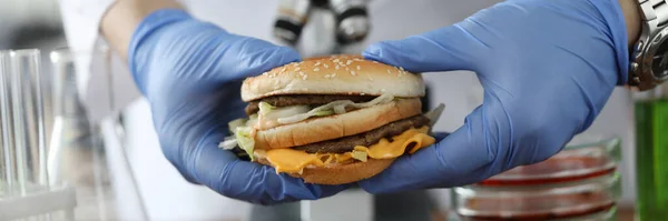 Cientista em luvas de borracha segurar hambúrguer na frente de microscópio e tubos de ensaio de vidro em close-up laboratório químico — Fotografia de Stock