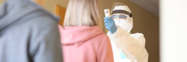 Klinikte kuyruktaki insanların sıcaklığını ölçen koruyucu tulumlu bir doktor.