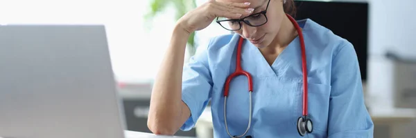 Jeune femme médecin assis à table et tenant la tête devant l'ordinateur portable dans le bureau — Photo