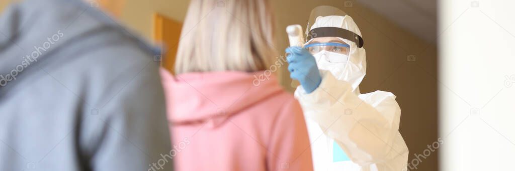 Doctor in protective overalls measuring temperature of people in line at clinic