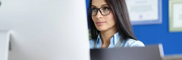Mujer con gafas y camisa azul sentarse en el lugar de trabajo y mirar a la pantalla del ordenador. — Foto de Stock