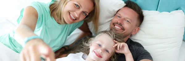 Klein kind meisje met je ouders liggen op bed portret — Stockfoto