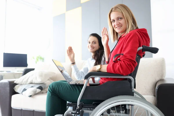 Жінка в інвалідному візку з ноутбуком на колінах махає в привітання з дівчиною — стокове фото