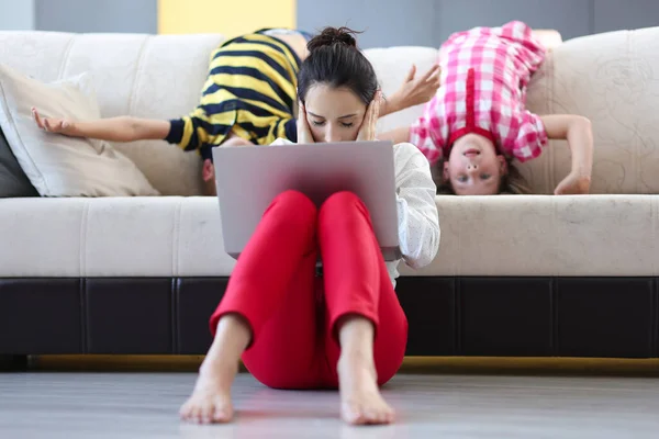 Tired woman with laptop sits on floor with closed palms ears behind on couch children indulge — Stock Photo, Image