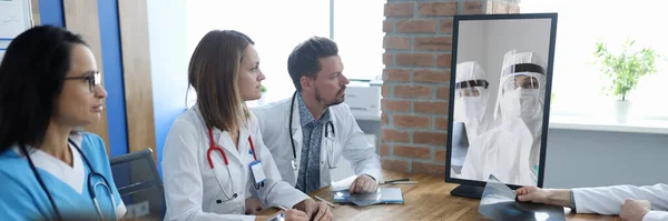 Doctors at meeting communicating via video communication with colleagues in protective overalls