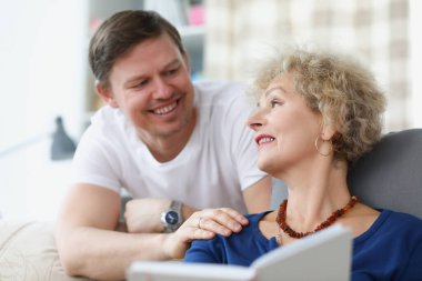 Yaşlı bir kadın kanepede oturuyor ve genç adama gülümsüyor.