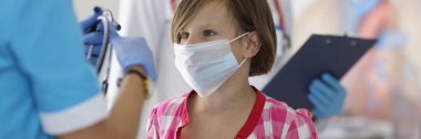 Koruyucu tıbbi maskeli küçük kız doktorun yanında duruyor.