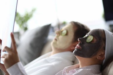 Genç adam ve kadın nemlendirici yüz maskeleriyle kanepede yatıyorlar.