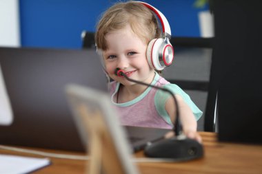 Çalışma masasında, bilgisayarı kulaklıklı, gülümseyen küçük bir kız mikrofonun yanında duruyor.