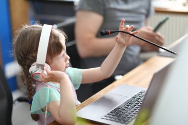 Kulaklıklı küçük kız mikrofonun önünde dizüstü bilgisayarın yanında duruyor.