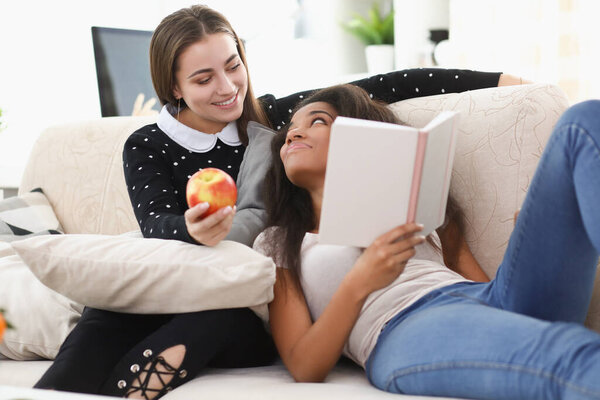 Two young girls are sitting on couch and reading book at home