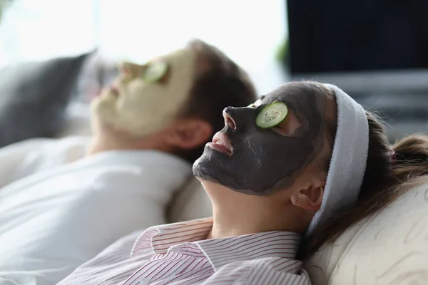 Jovem homem e mulher deitados com máscaras cosméticas em seus rostos e fatias de pepino em seus olhos — Fotografia de Stock