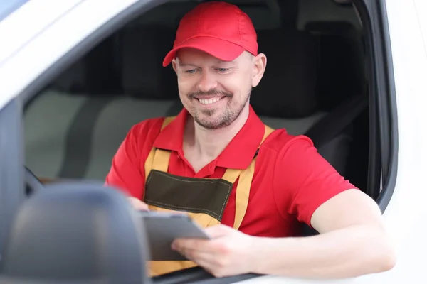 Man courier in uniform sitting at wheel of car with digital tablet in his hands — Stock Photo, Image