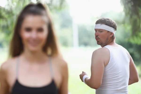 Товстий чоловік у спортивному одязі дивиться на молоду дівчину, що бігає в парку — стокове фото