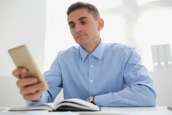 Young man in blue shirt looks at smartphone screen while sitting at his desk