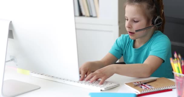 Klein meisje in hoofdtelefoon typt op computer toetsenbord en spreken in microfoon 4k film — Stockvideo