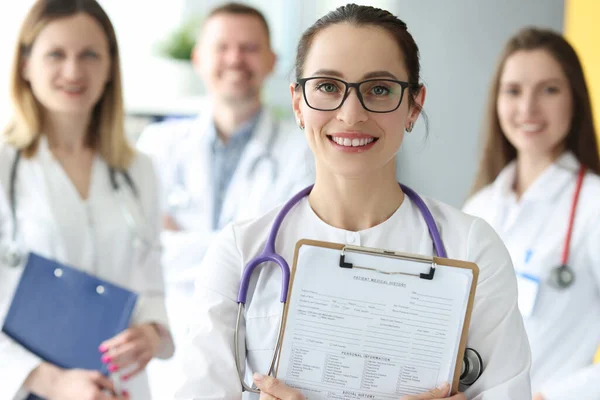 Porträtt av kvinnlig läkare i glasögon med patient sjukdomshistoria i händerna på bakgrund av kollegor — Stockfoto