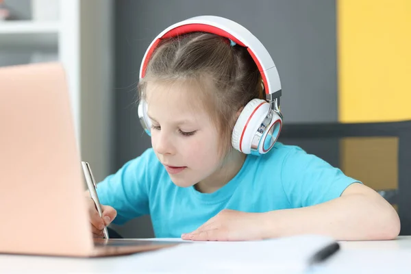Κορίτσι με ακουστικά κάνει σημειώσεις στο σημειωματάριο στο τραπέζι είναι φορητό υπολογιστή — Φωτογραφία Αρχείου