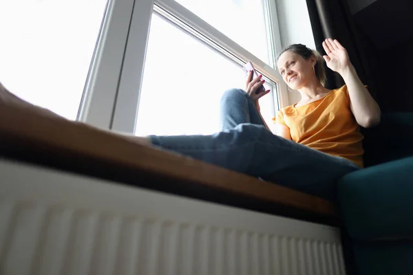 Молода жінка сидить на підвіконні і махає рукою на екрані мобільного телефону — стокове фото