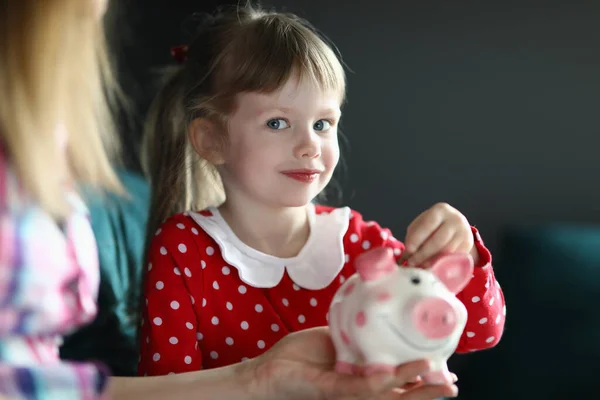 Menina com sua mãe joga moeda no banco rosa piggy — Fotografia de Stock