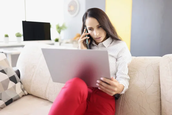 Jovem mulher falando no telefone e trabalhando no laptop enquanto está sentado no sofá — Fotografia de Stock