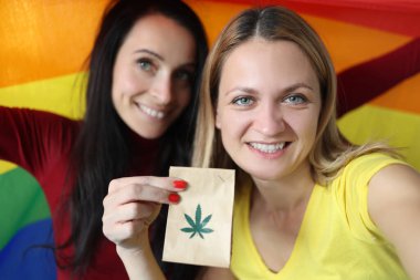 İki gülümseyen kadın ellerinde lgbt bayrağı ve bir paket marihuana tutuyor.