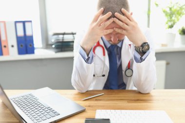 Yorgun doktor masada oturuyor ve kafasını tutuyor.