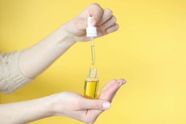 Sarı arka planda bir şişe kozmetik yağı tutan kadın elleri.