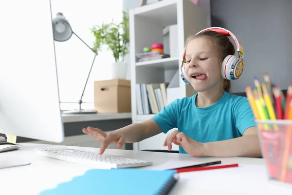 Kleines Mädchen mit Kopfhörern sitzt am Tisch und tippt auf Computertastatur — Stockfoto