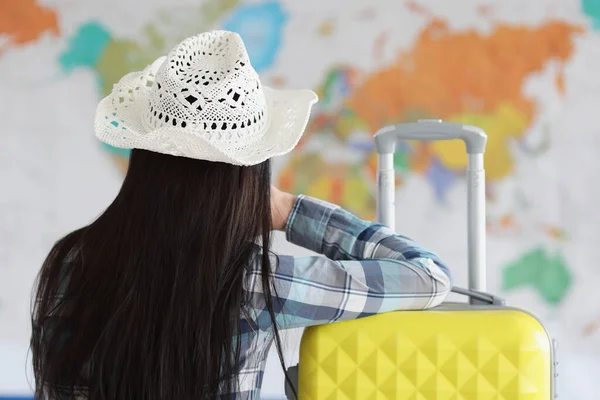 Woman sits with her back in white hat and suitcase and looks at map of world — Stock Photo, Image