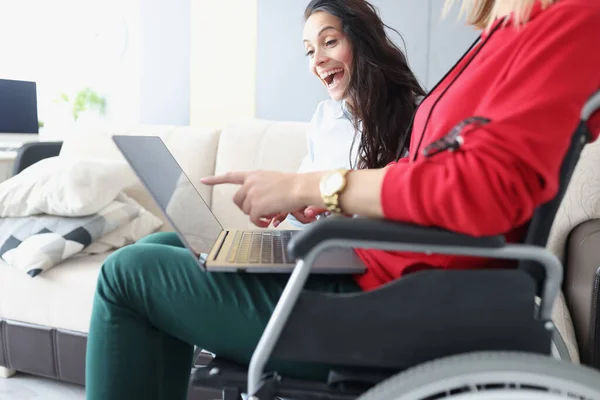 Інвалідність жінка в інвалідному візку з ноутбуком на колінах показує її радісну подругу її досягнення в роботі — стокове фото