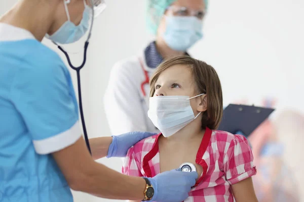 Dokter bertopeng medis pelindung mendengarkan hati dengan stetoskop untuk gadis kecil — Stok Foto