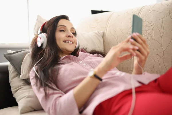Jovem encontra-se no sofá usando fones de ouvido e olha para o smartphone — Fotografia de Stock