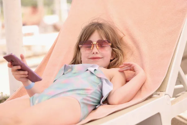 Pequeña chica sonriente en gafas de sol se encuentra en la tumbona con teléfono inteligente en sus manos — Foto de Stock