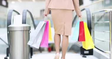 Alışveriş merkezinde yürüyen merdivene binen takım elbiseli bir kadının arkası. Elinde bir sürü renkli çanta var. 4K film.