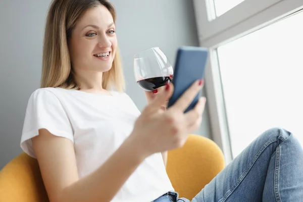 Молода жінка тримає мобільний телефон в руках і п'є червоне вино зі скла вдома — стокове фото