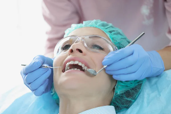 Tandarts onderzoeken tanden van vrouw patiënt in beschermende bril en pet met behulp van tandheelkundige hulpmiddelen — Stockfoto